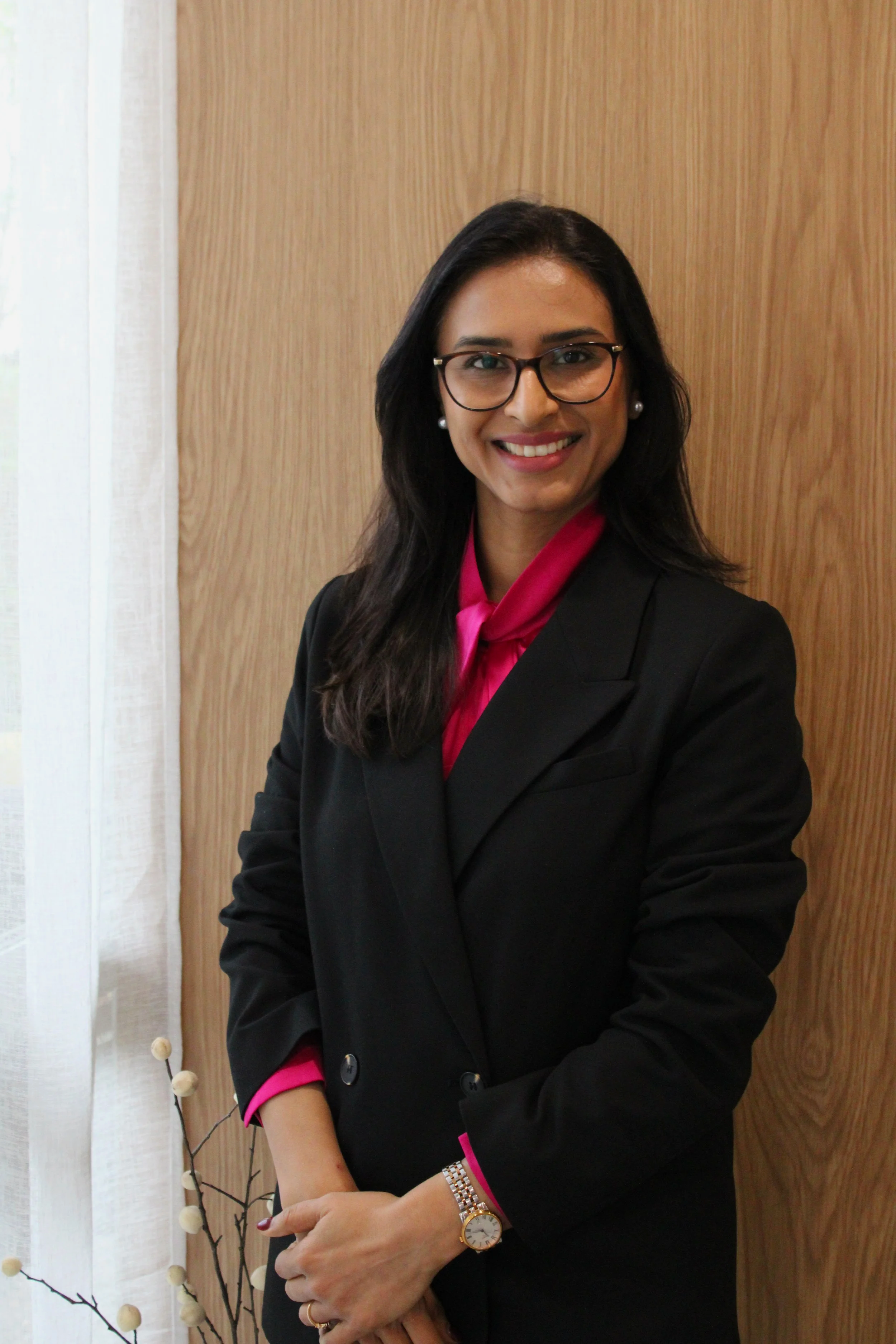 Professional headshot of a dermatologist with long dark hair, wearing a navy pinstripe blazer over a floral blouse, smiling against a neutral grey background.