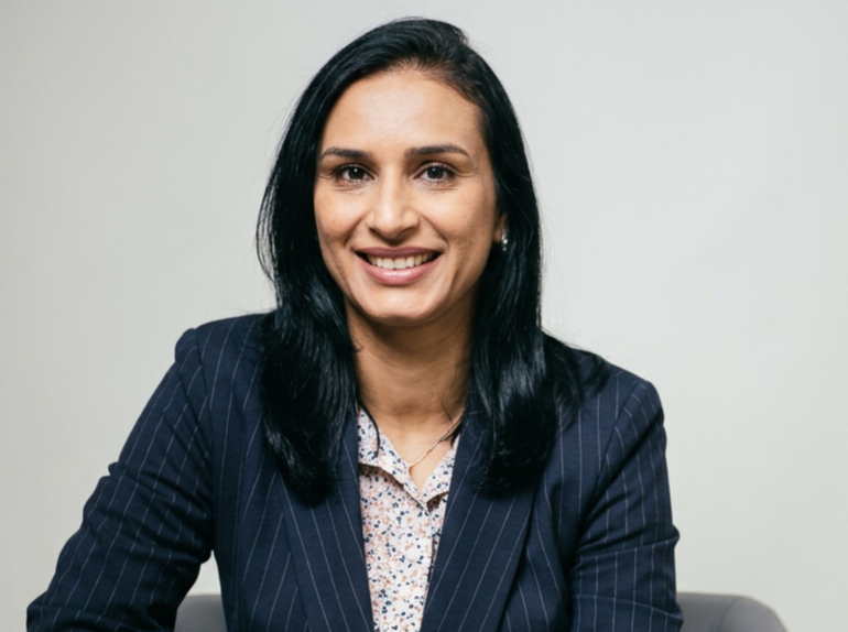 Professional headshot of a dermatologist with long dark hair, wearing a navy pinstripe blazer over a floral blouse, smiling against a neutral grey background.