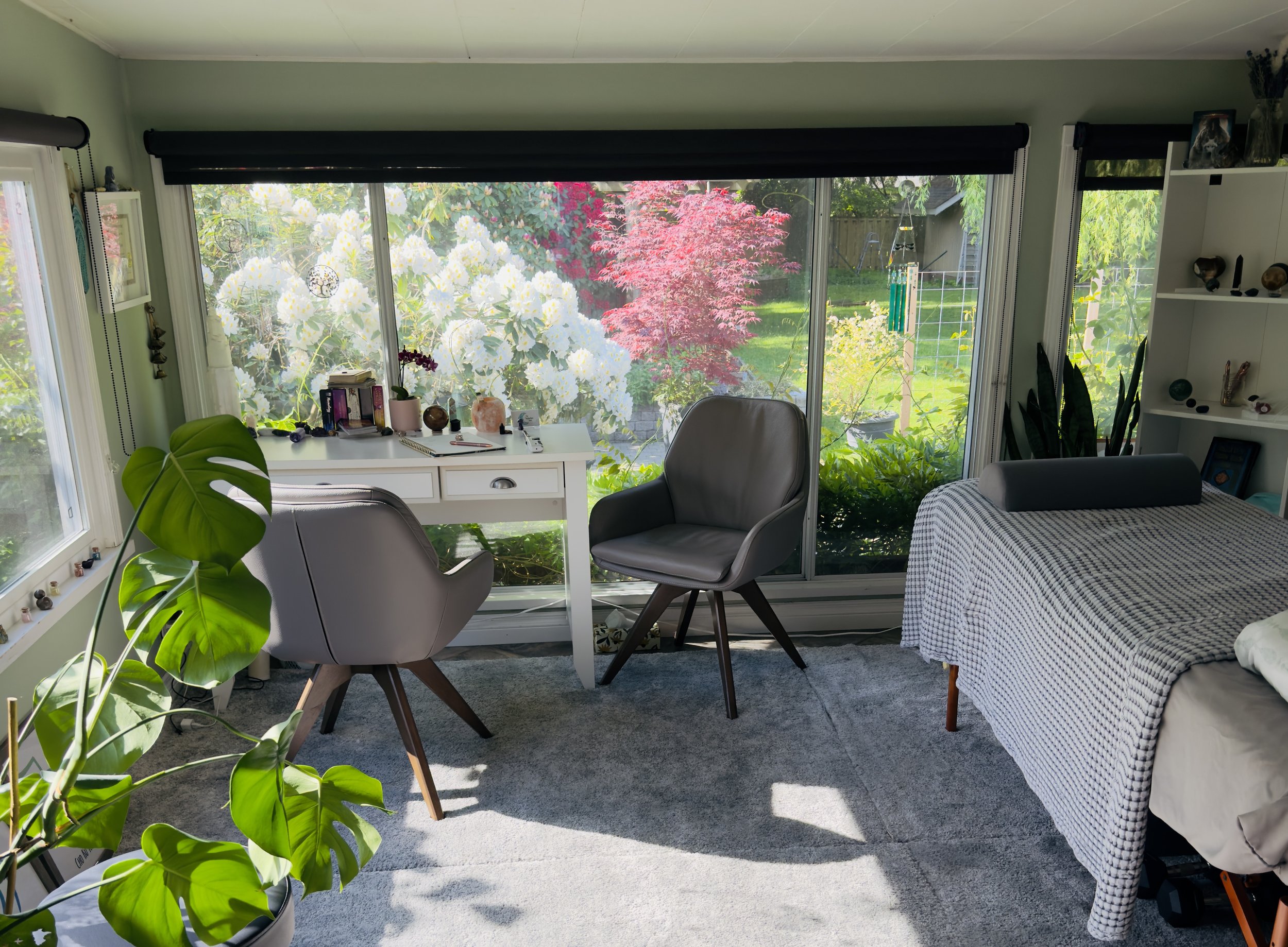 Living room with large windows overlooking a garden with white flowers and pinkish-red trees. Contains a white desk with items, two gray chairs, a bed with a checkered blanket, a white shelf with decorative items, a large green plant, and a gray carpet.