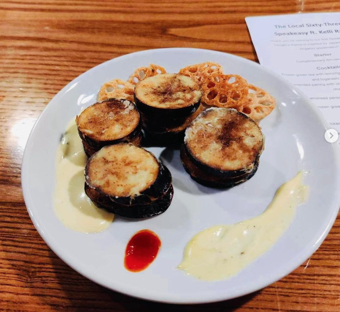 A plate with cooked eggplant slices, lotus root chips, a small dollop of red sauce, and dollops of yellowish-white sauce, on a wooden table.