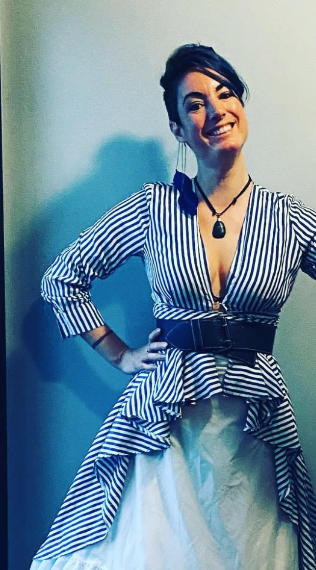 Woman smiling, wearing a striped top with ruffles, a wide belt, and a white skirt, standing in front of a plain wall.