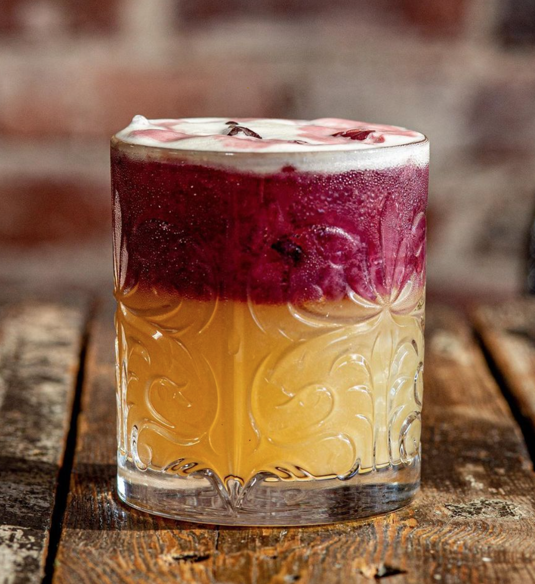 A glass of layered beverage with a dark red top layer and a yellow bottom layer, garnished with ice and condensation on the glass, placed on a wooden surface with a blurred rustic background.