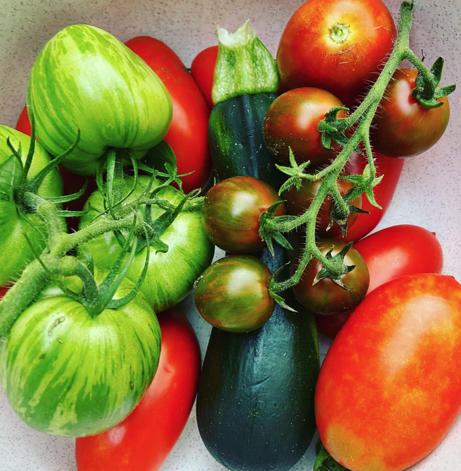 Assorted fresh vegetables including heirloom tomatoes, cucumbers, and green and red peppers on a light-colored surface.