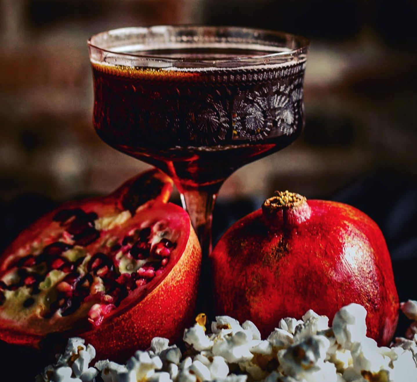 A glass of red wine, a halved pomegranate with seeds visible, a whole pomegranate, and scattered popcorn on a dark surface.