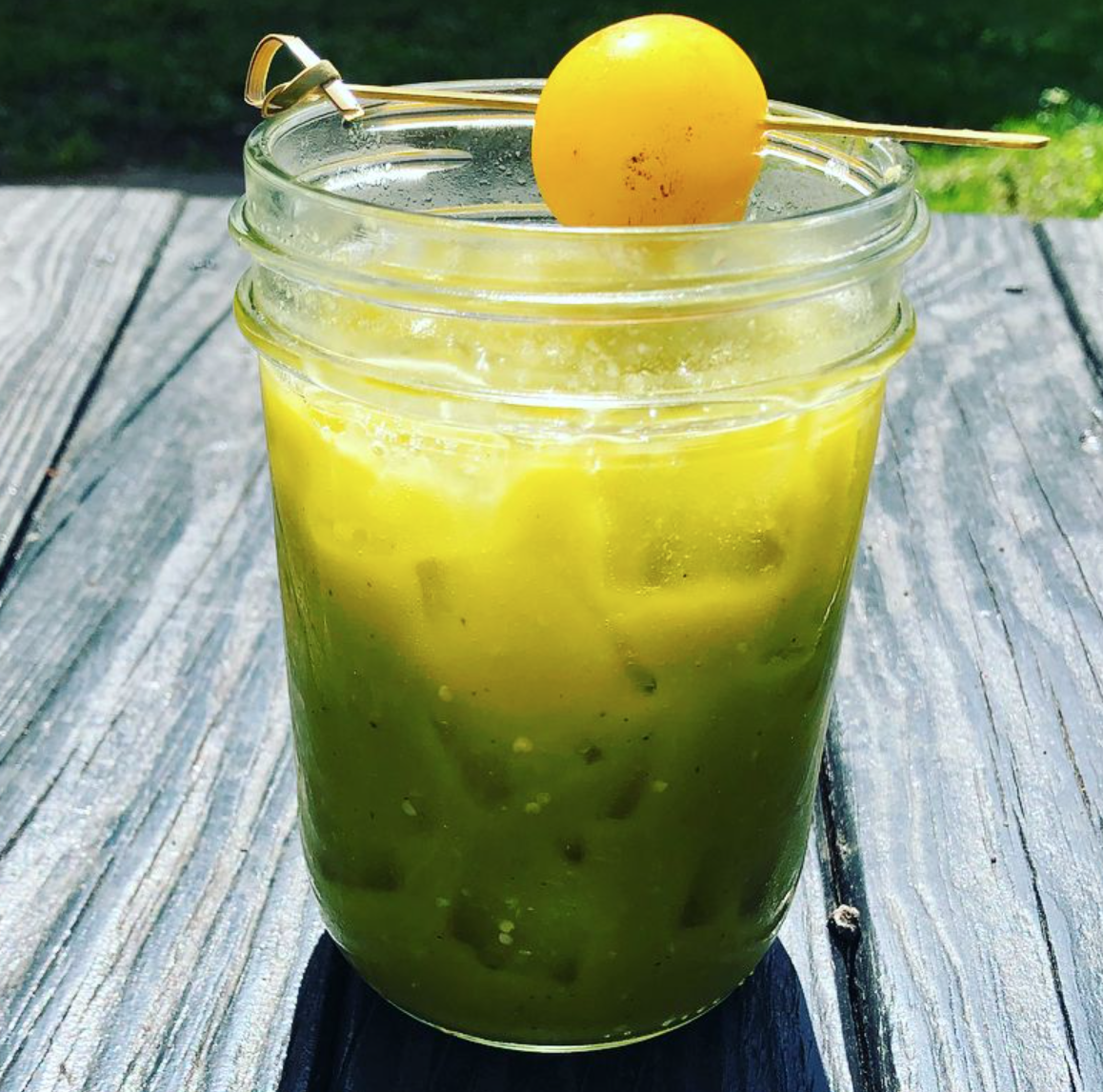 A mason jar filled with green and yellow slushy drink, garnished with a green olive on a skewer, on a weathered wooden table outdoors.