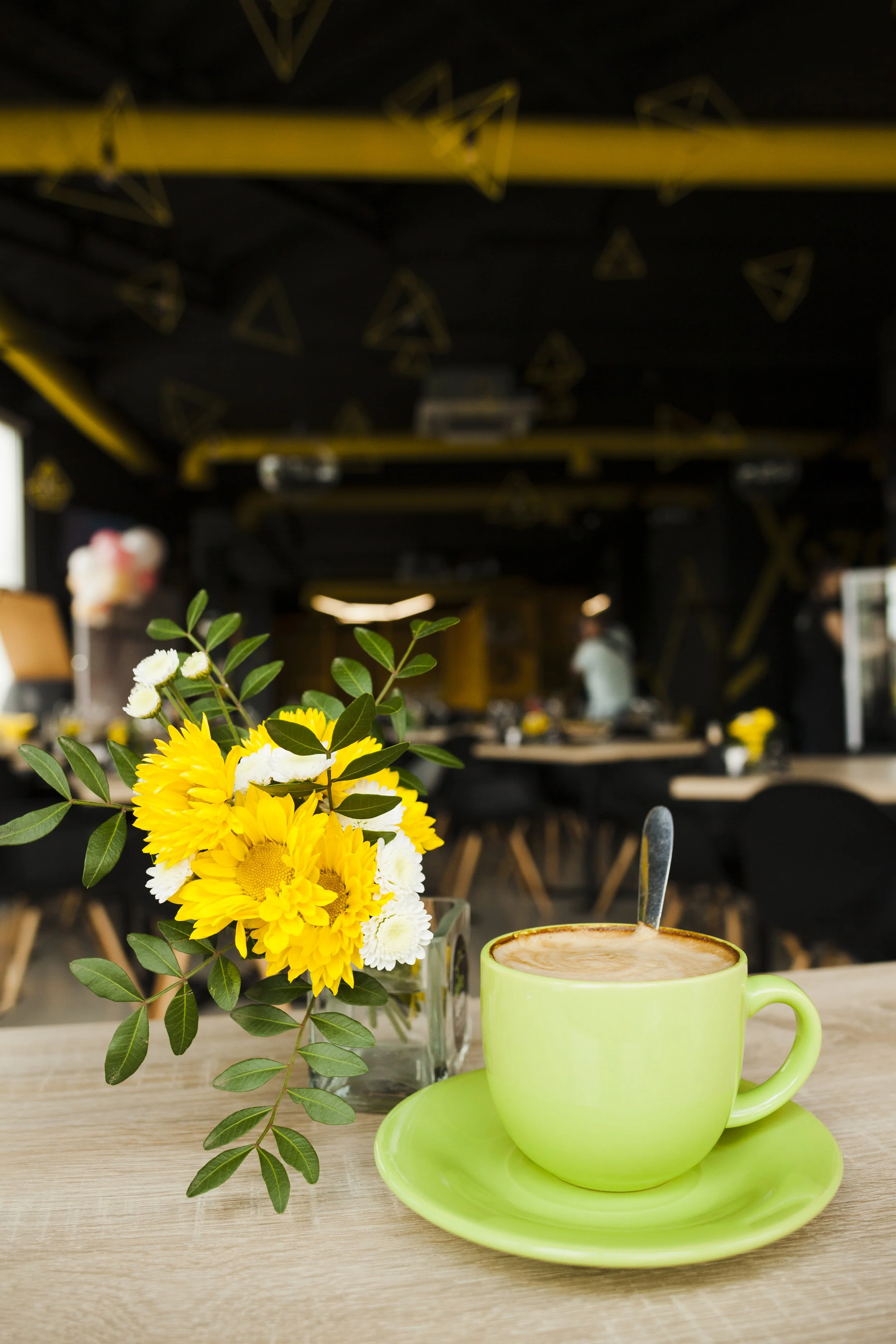 A bright yellow flower bouquet with green leaves in a clear jar, a green coffee mug with a hot beverage, and a matching green saucer on a wooden table in a cafe with black walls and yellow triangle decals.