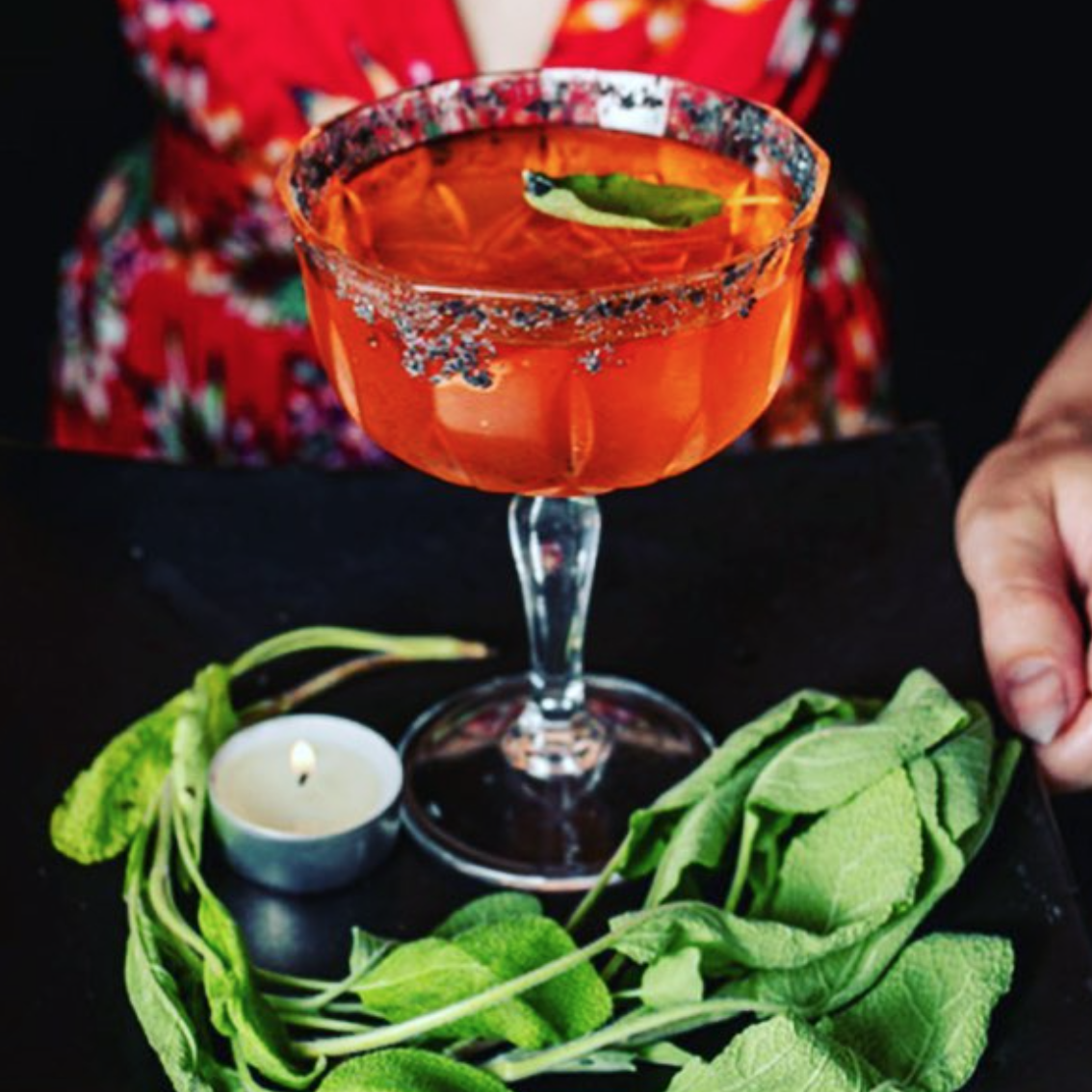 A close-up of a pink cocktail in a glass with a salted rim, garnished with a green chili, on a black surface with fresh green herbs and a small lit candle nearby.