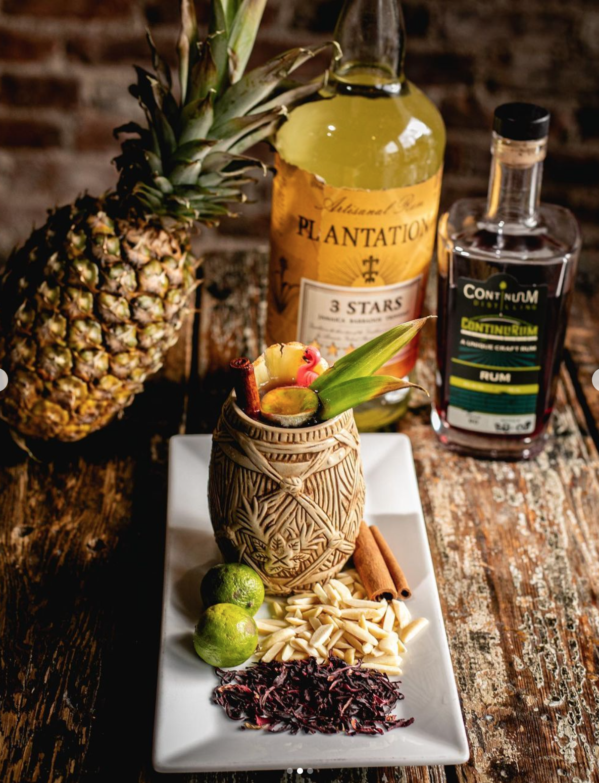 A tropical cocktail garnished with pineapple, lime, and cinnamon stick on a decorative ceramic mug, accompanied by two limes, white chocolate, dried hibiscus, and cinnamon sticks on a white rectangular plate, with pineapple and liquor bottles in the 