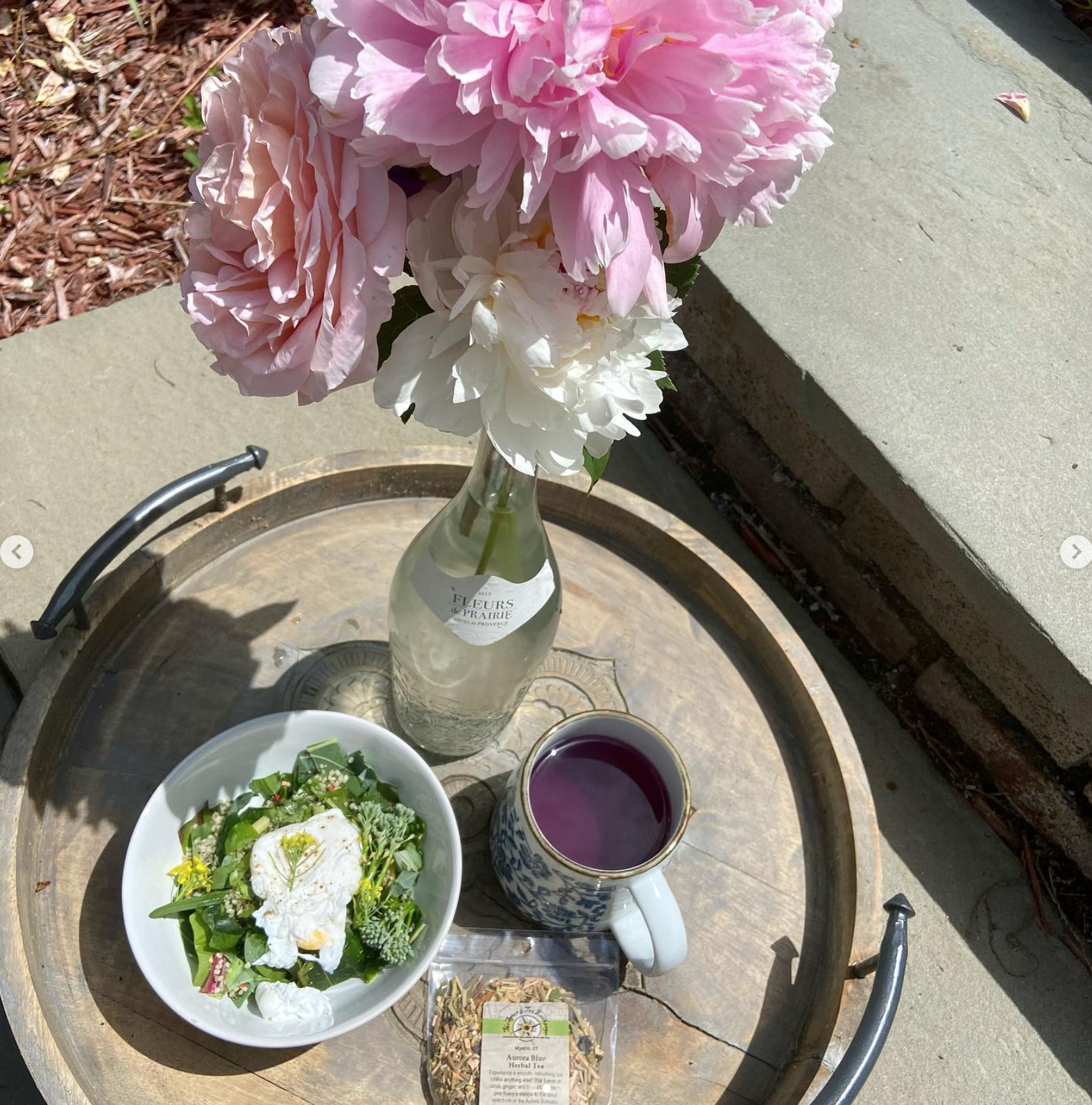 A tray with a vase of pink and white peonies, a bowl of salad with a poached egg, a mug of purple herbal tea, a bottle of water, and a packet of herbal tea on a concrete surface outdoors.