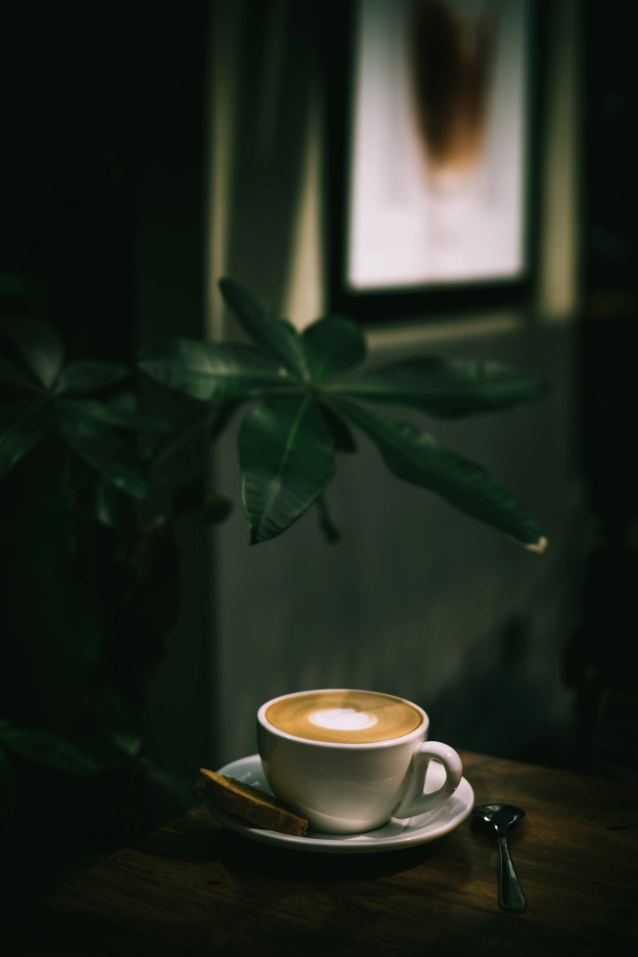 A white cup of cappuccino with foam art on top, placed on a white saucer with a cookie and spoon, on a wooden table. Green houseplants and a framed picture are in the background.