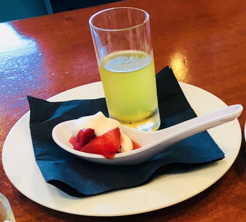 A glass of yellow beverage and a small dish with strawberries and cream on a white plate with a black napkin.