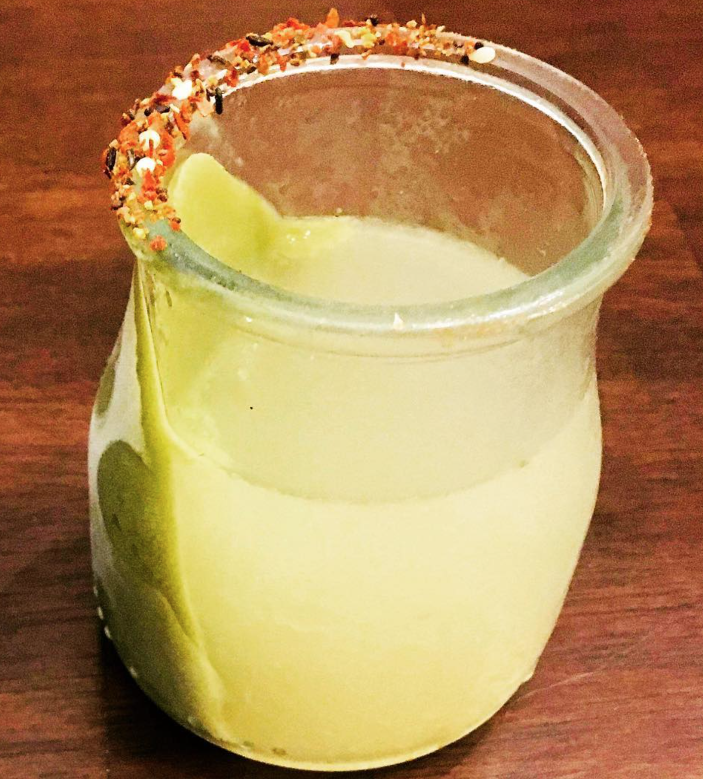 A mason jar with a lime wedge and a salted rim with chili powder and spices, filled with a yellow drink, on a wooden surface.