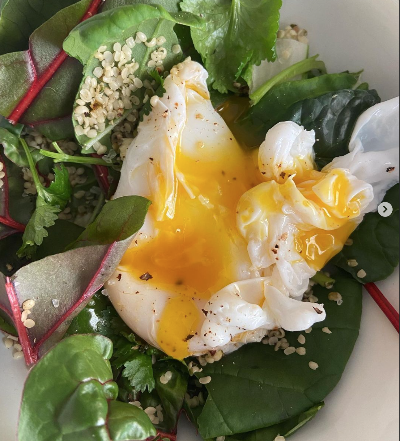 Poached eggs with runny yolk on a bed of mixed greens, including spinach, lettuce, and herbs, garnished with sesame seeds.