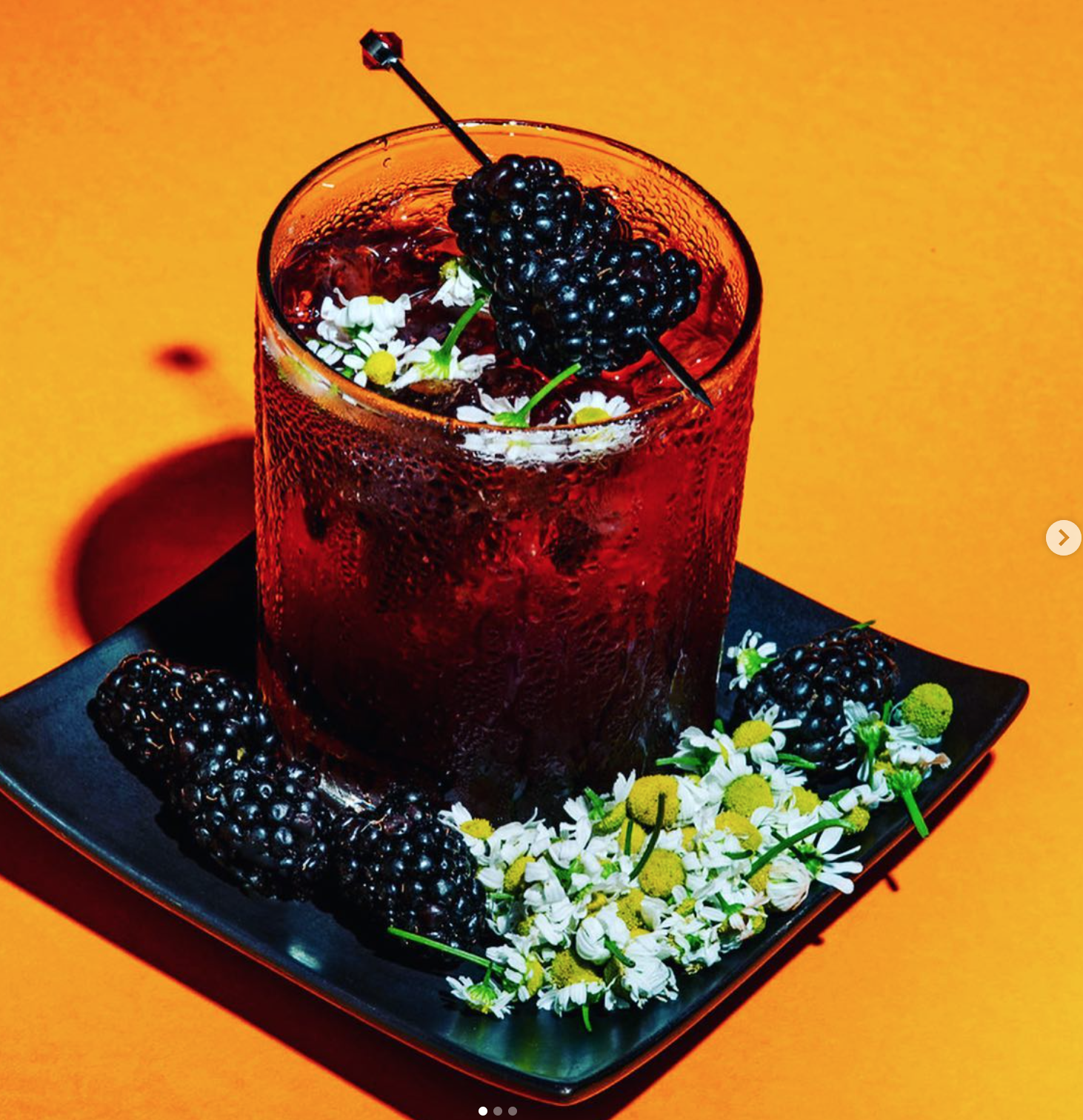 A glass of dark red berry cocktail garnished with blackberries on top, alongside blackberries and white flowers with yellow centers on a black square plate, set against a yellow background.