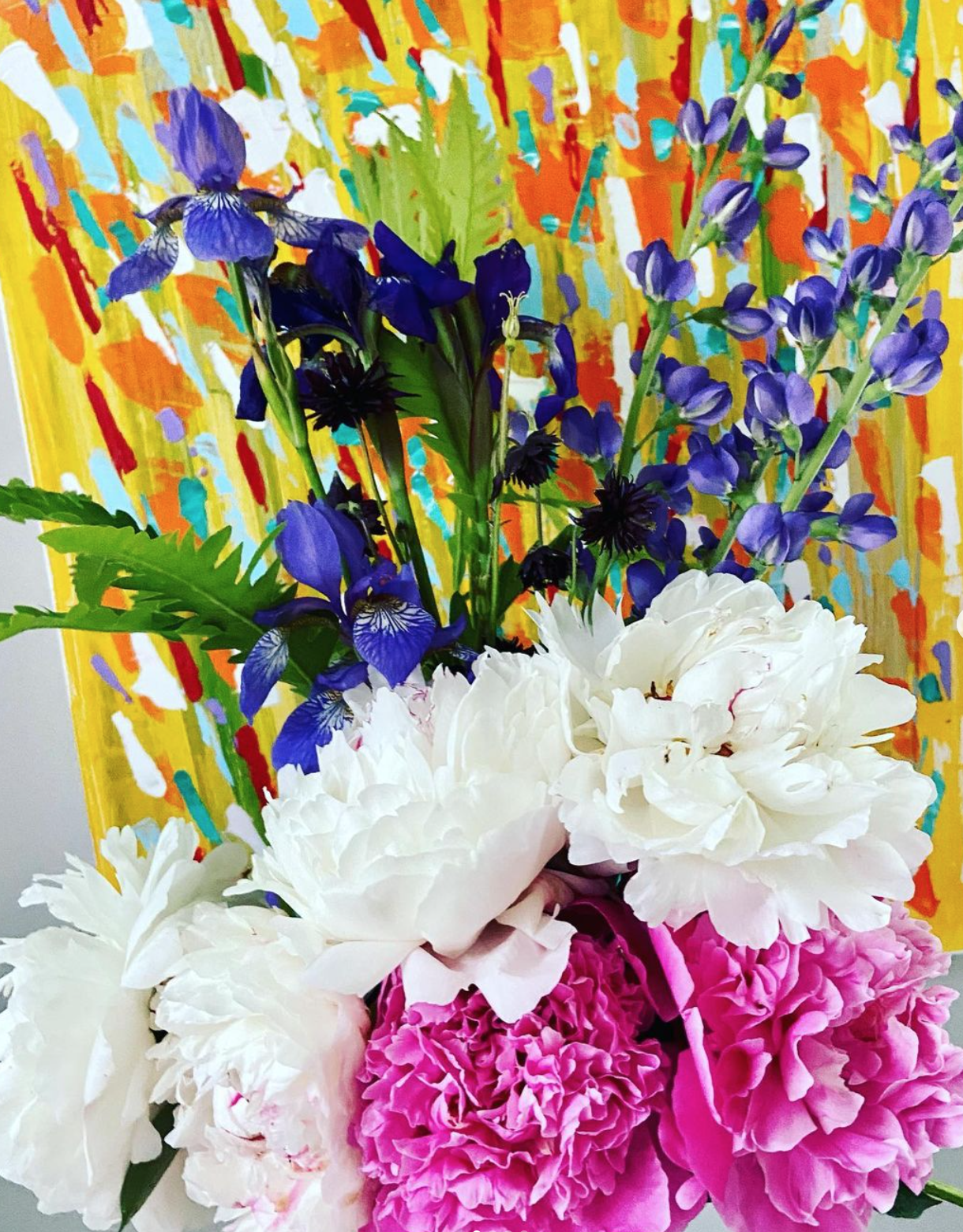 A colorful floral arrangement with purple, white, and pink peonies and purple irises set against a vibrant abstract yellow, orange, green, red, blue, and purple background.