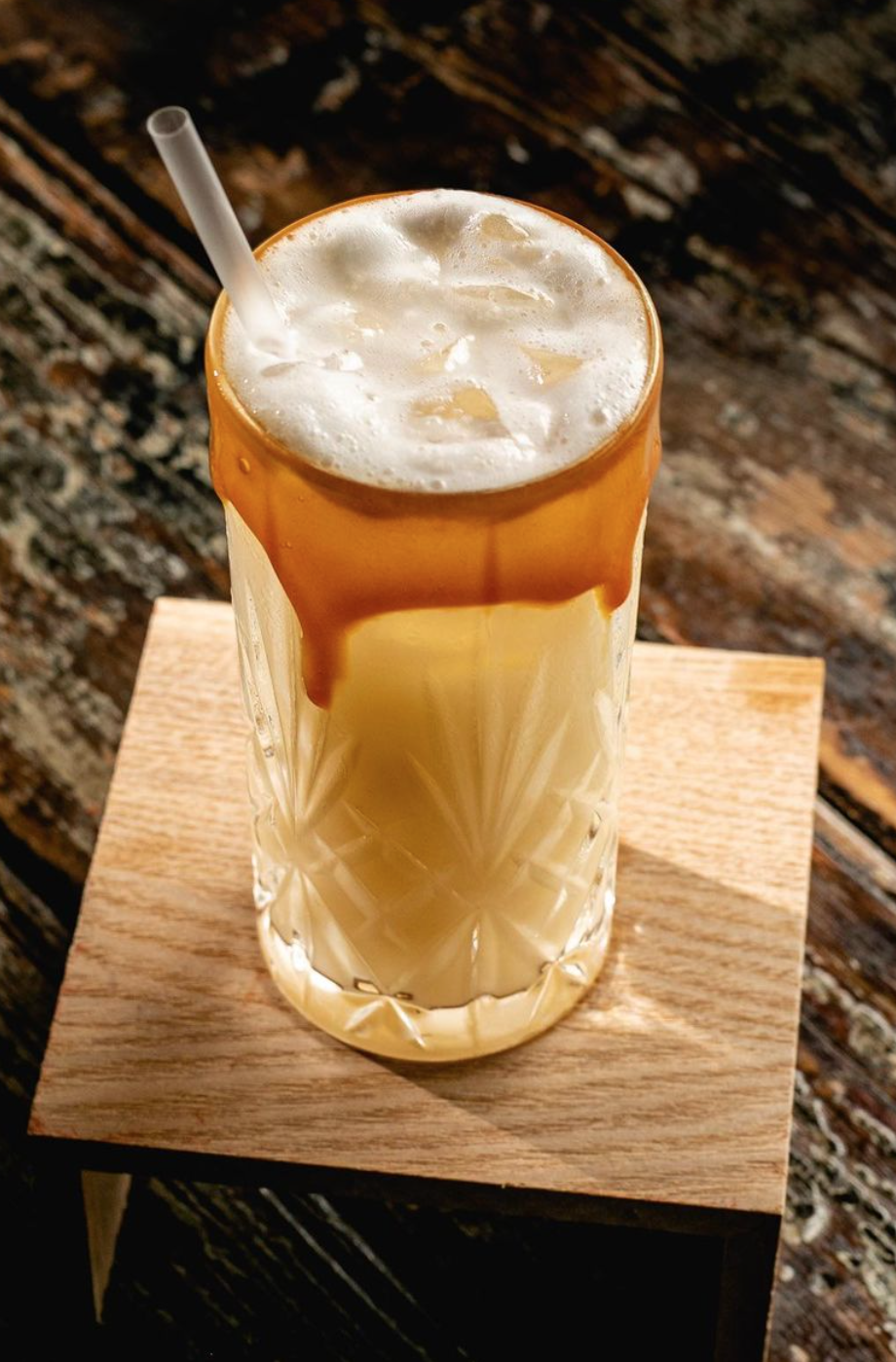 A tall glass of iced coffee topped with cream and caramel syrup, served with a straw on a wooden coaster.
