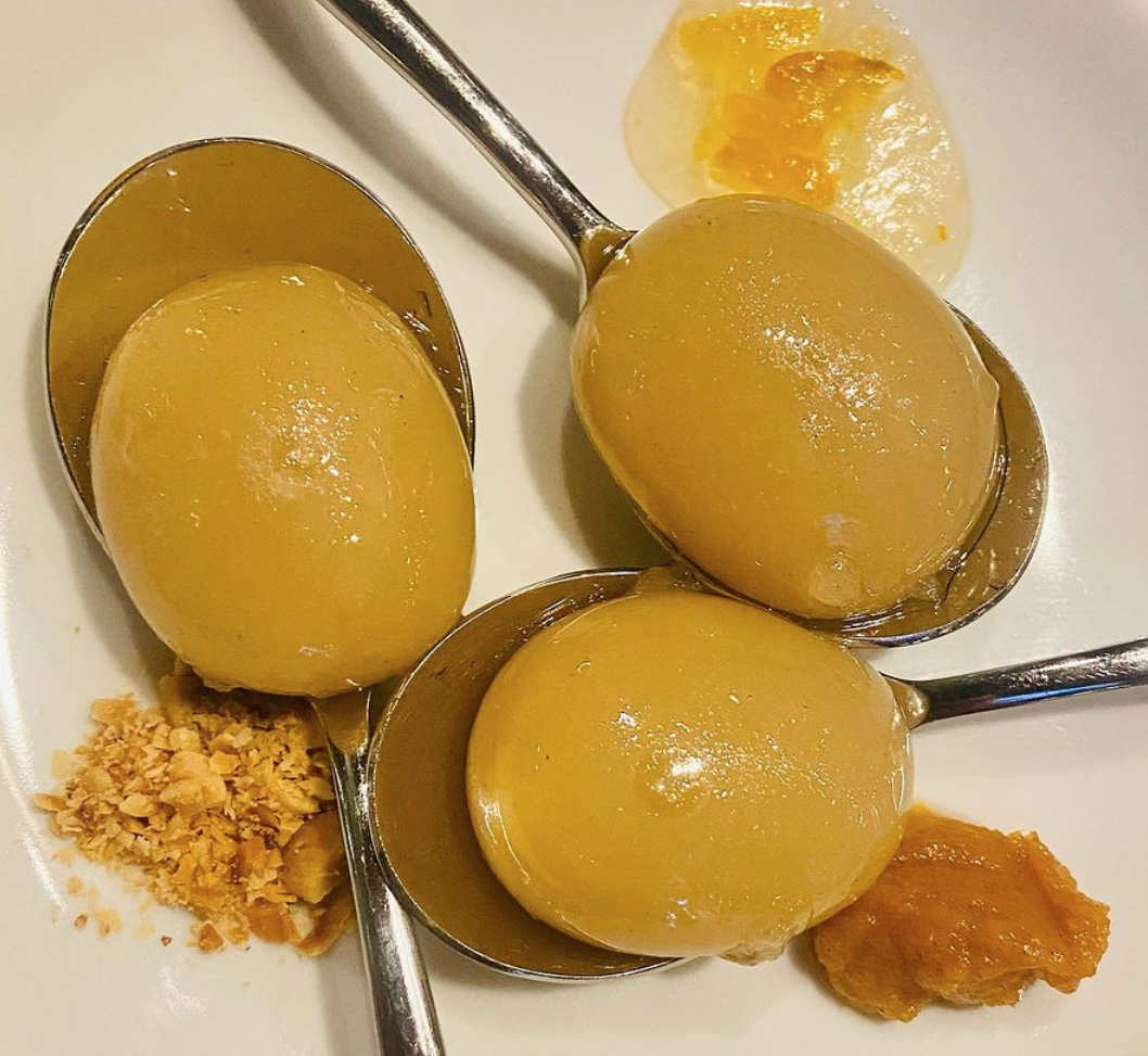 Four metal spoons filled with jellied yellow-green eggs, garnished with crushed nuts, mashed banana, and slices of banana with honey on a white plate.