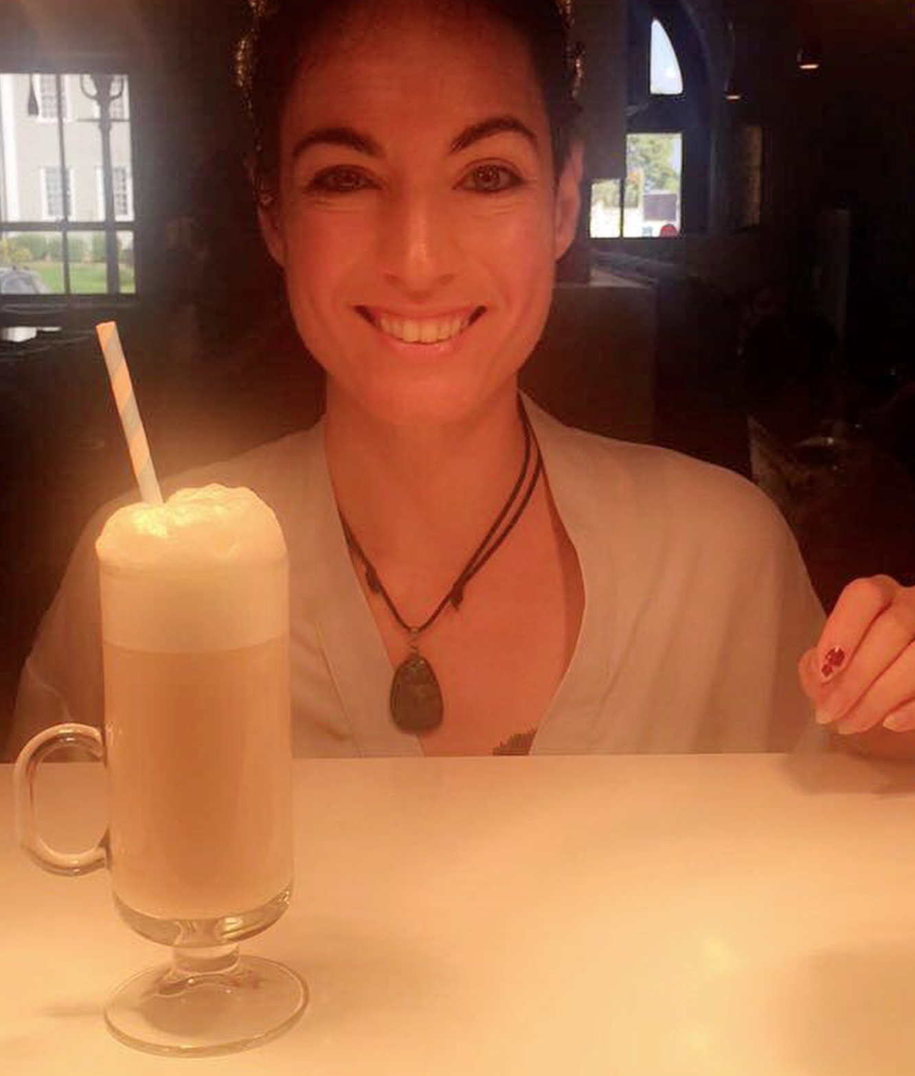 A woman with short dark hair smiling at a table with a large milkshake topped with whipped cream and a straw in a dimly lit restaurant.