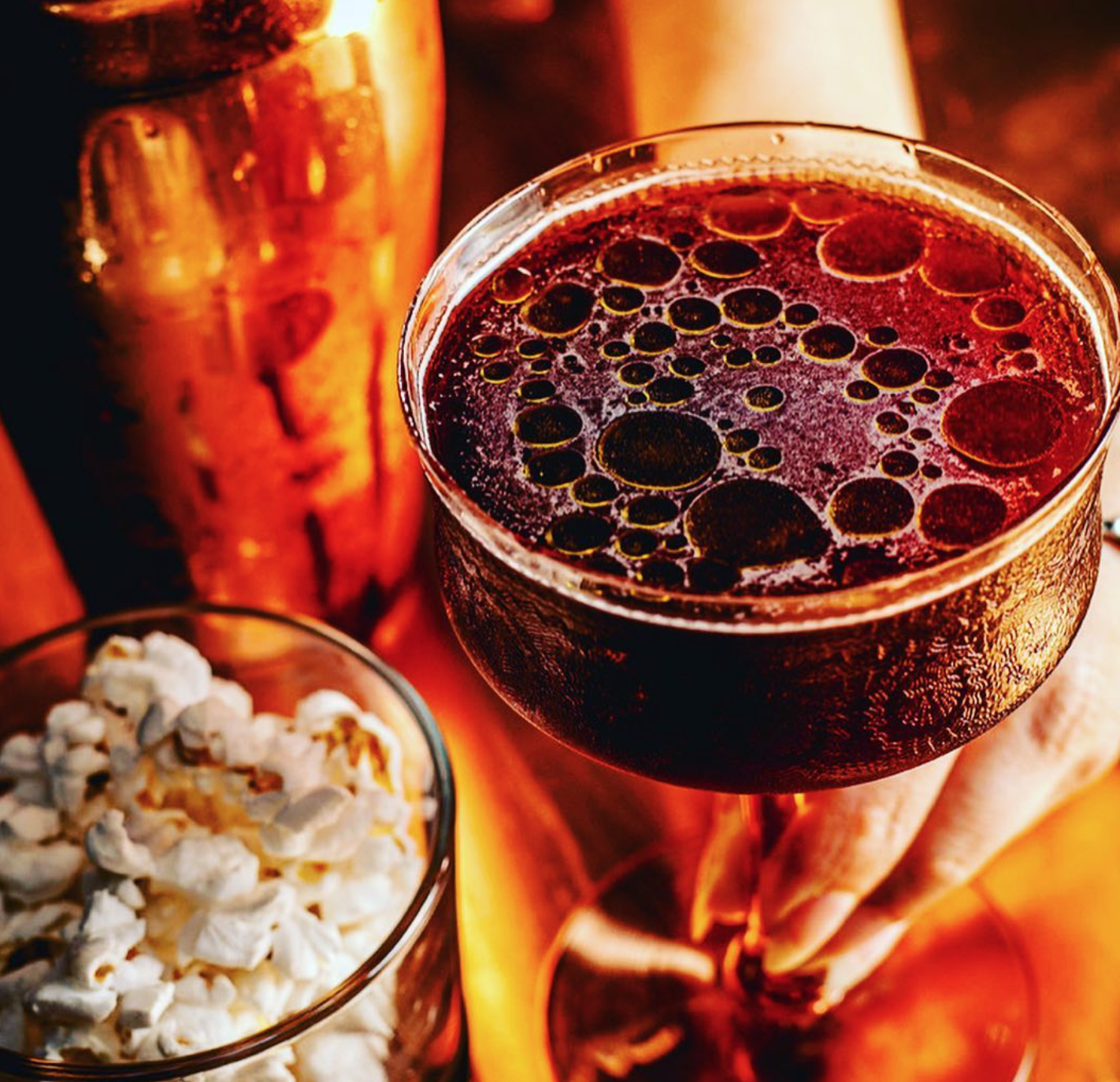 Close-up of a glass of dark soda with bubbles, a glass of beer, and a bowl of popcorn on a warm-colored table.
