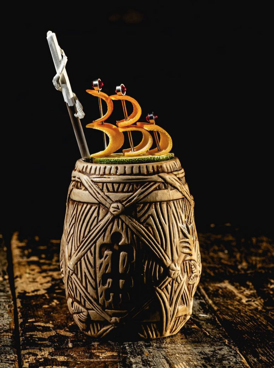 A tiki-style mug with decorative carved designs, topped with yellow fruit slices shaped like sailboats, and small decorative swords, with a razor blade inserted in the fruit sailboat.