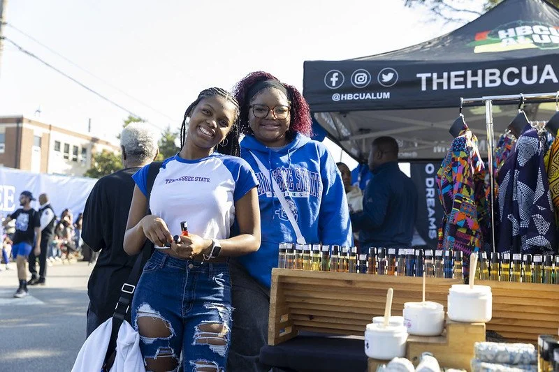 Two young women smiling at an outdoor event, standing behind a table with small bottles, near a booth with a tent labeled 'THEHBCUALUM' and colorful clothing racks.