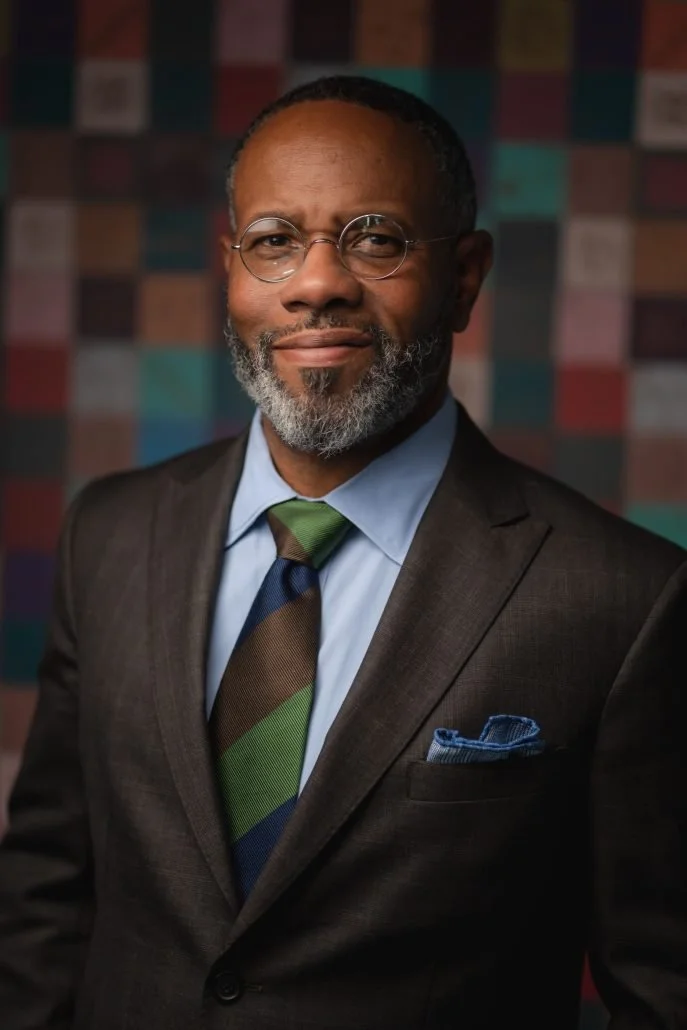 A professional man with glasses, a gray beard, and a warm expression, wearing a dark suit, light blue shirt, and a multicolored striped tie, standing against a blurred multicolored background.