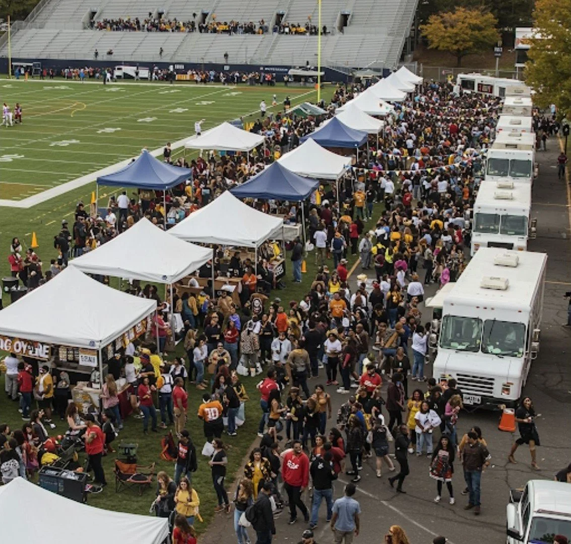 A crowded football field with tents and food trucks set up along the sideline, where people are gathered for an event or celebration.