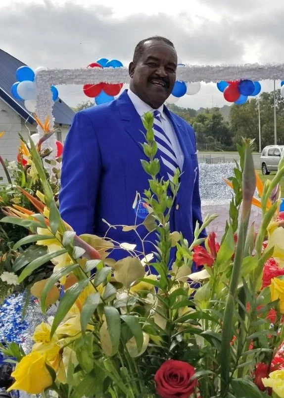 A man in a blue suit and striped tie standing behind a large bouquet of colorful flowers at an outdoor event decorated with red, white, and blue balloons and banners.