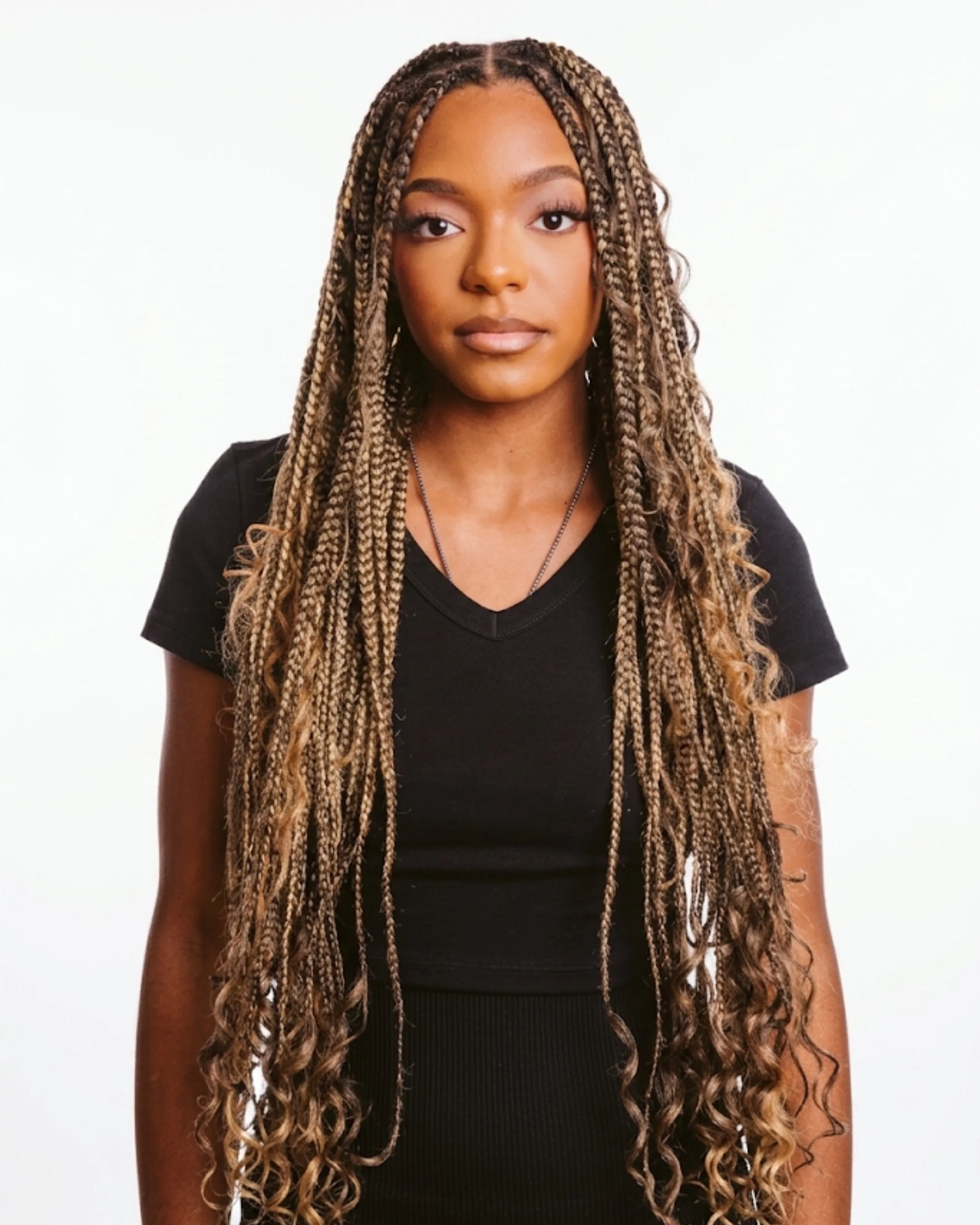 Portrait of a young woman with long, blonde-brown boho knotless braids, wearing a black top, against a plain white background.