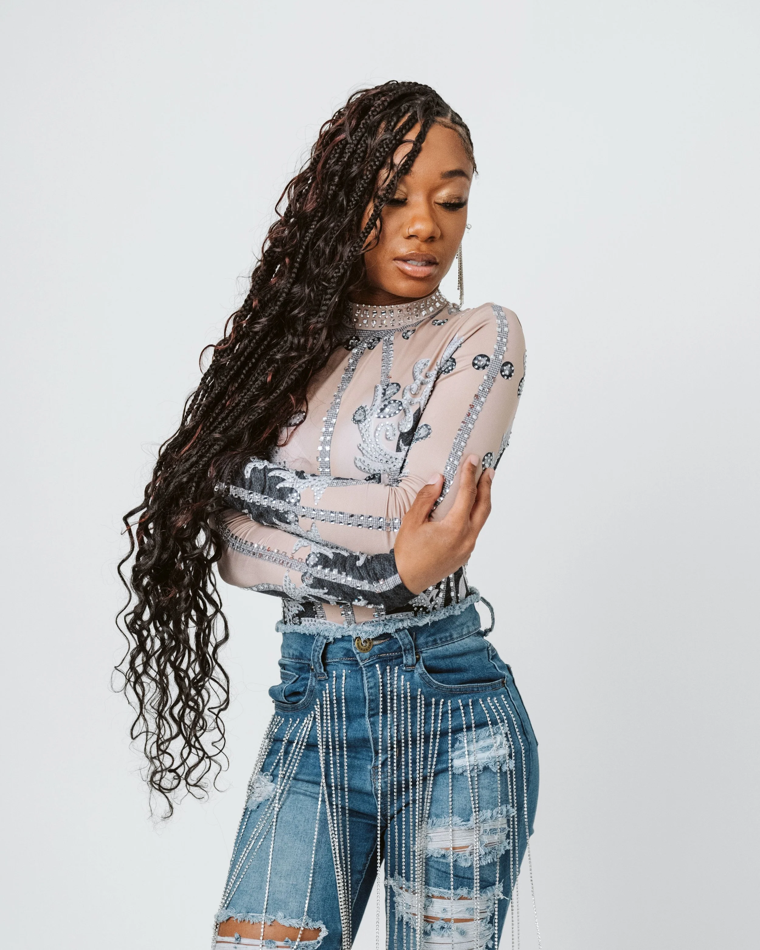 A woman with long boho feed in braids with curls and a tan complexion poses against a plain white background. She is wearing a sheer, with silver and black decorative patterns, and distressed blue jeans with silver embellishments.