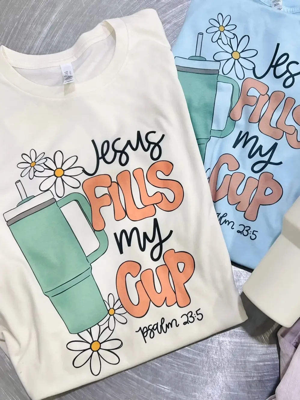 Two T-shirts with matching religious-themed designs are displayed on a table. The design includes a green mug with daisies, and the text 'Jesus fills my cup' with a biblical reference to Psalm 23:5.