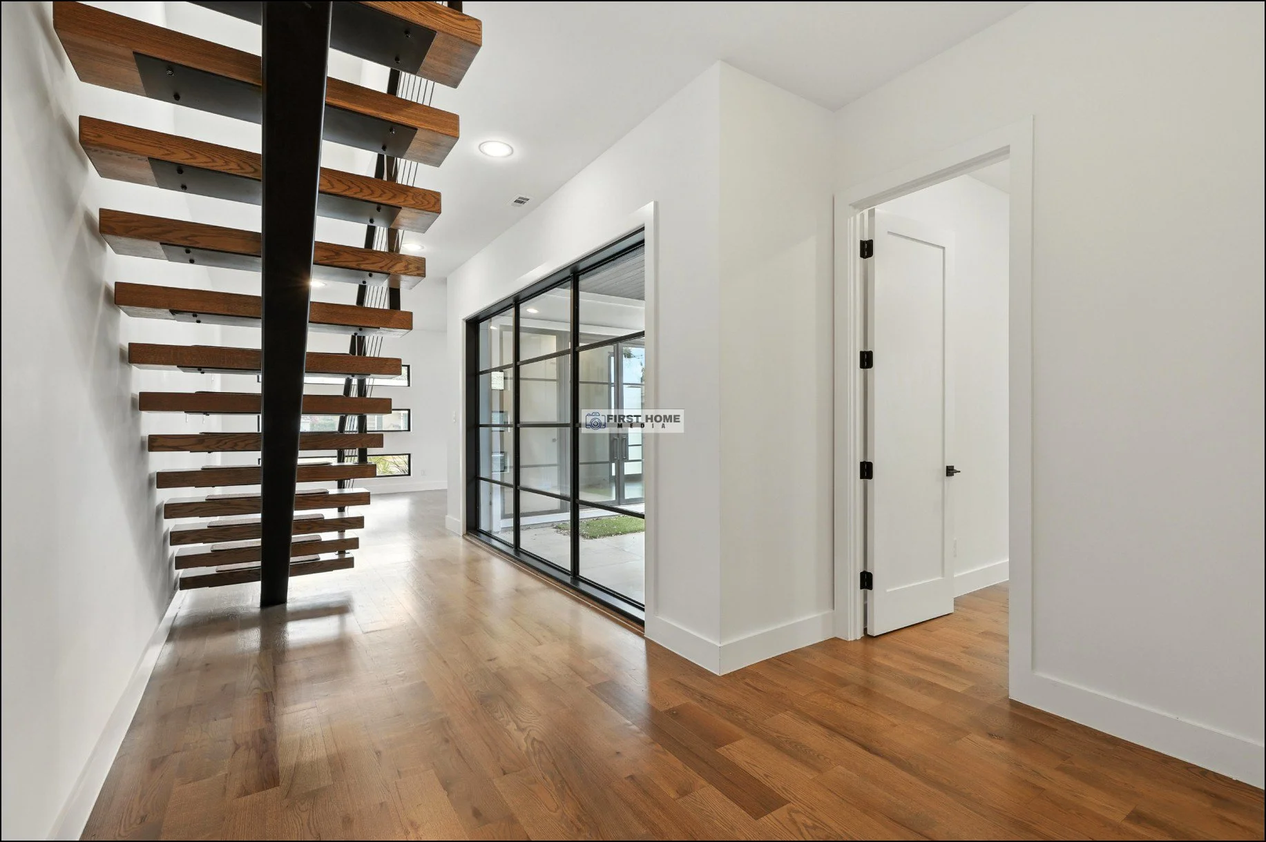 Interior view of a modern home with wooden floors, a staircase with open risers, a glass sliding door, and white walls.