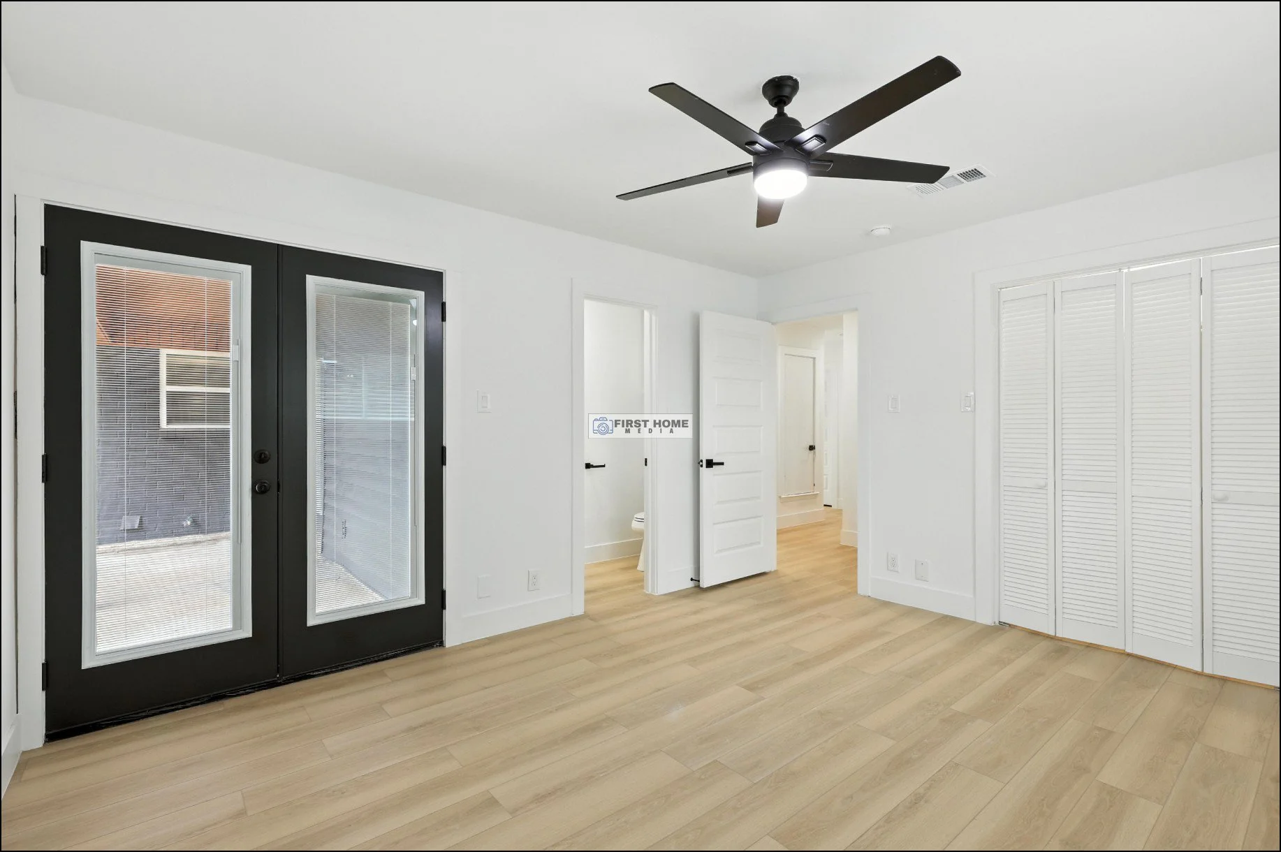 Empty room with white walls, wood flooring, black ceiling fan, black double doors with glass panes, and a white closet with louvered doors.