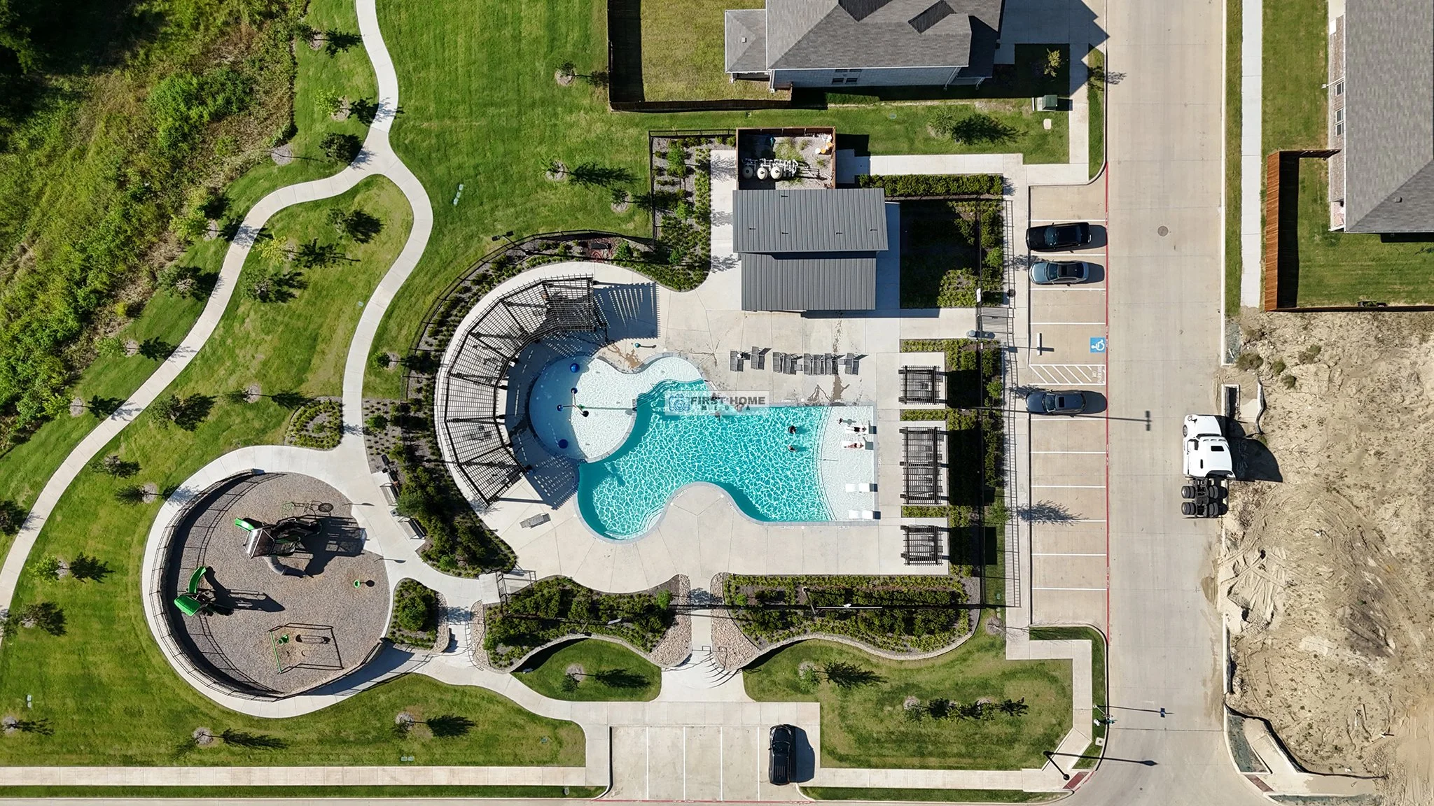 Aerial view of a community pool area with a water slide, splash pad, and swimming pool, surrounded by landscaped lawns, walking paths, parking lot, and neighboring houses.