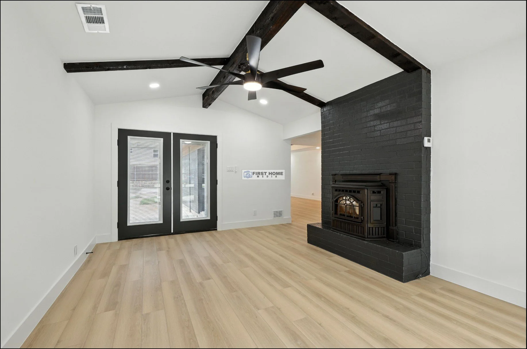 Living room with light wood flooring, white walls, black ceiling fan, black brick fireplace, and black glass double doors leading outside.