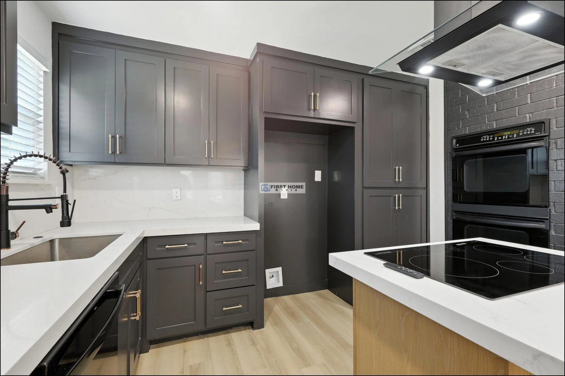 Modern kitchen with gray cabinets, white countertops, black appliances, and a black brick wall. Wood flooring and a window with blinds are also visible.