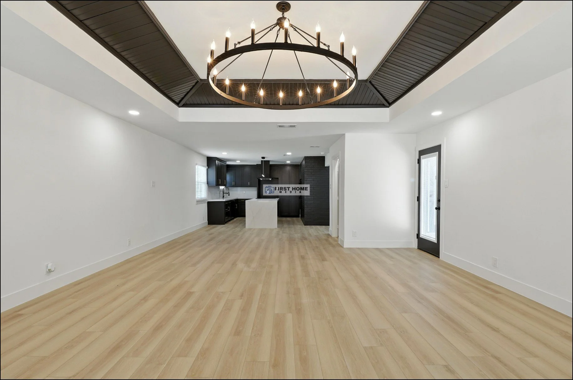 Empty open-concept living space with light wood flooring, white walls, black kitchen cabinets, and a chandelier in the ceiling.
