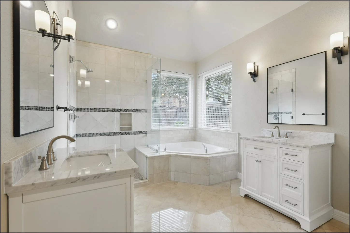 Modern bathroom with marble countertops, white cabinets, a corner bathtub, glass shower, and large mirrors. The walls are tiled, and there are stylish light fixtures above the mirrors.