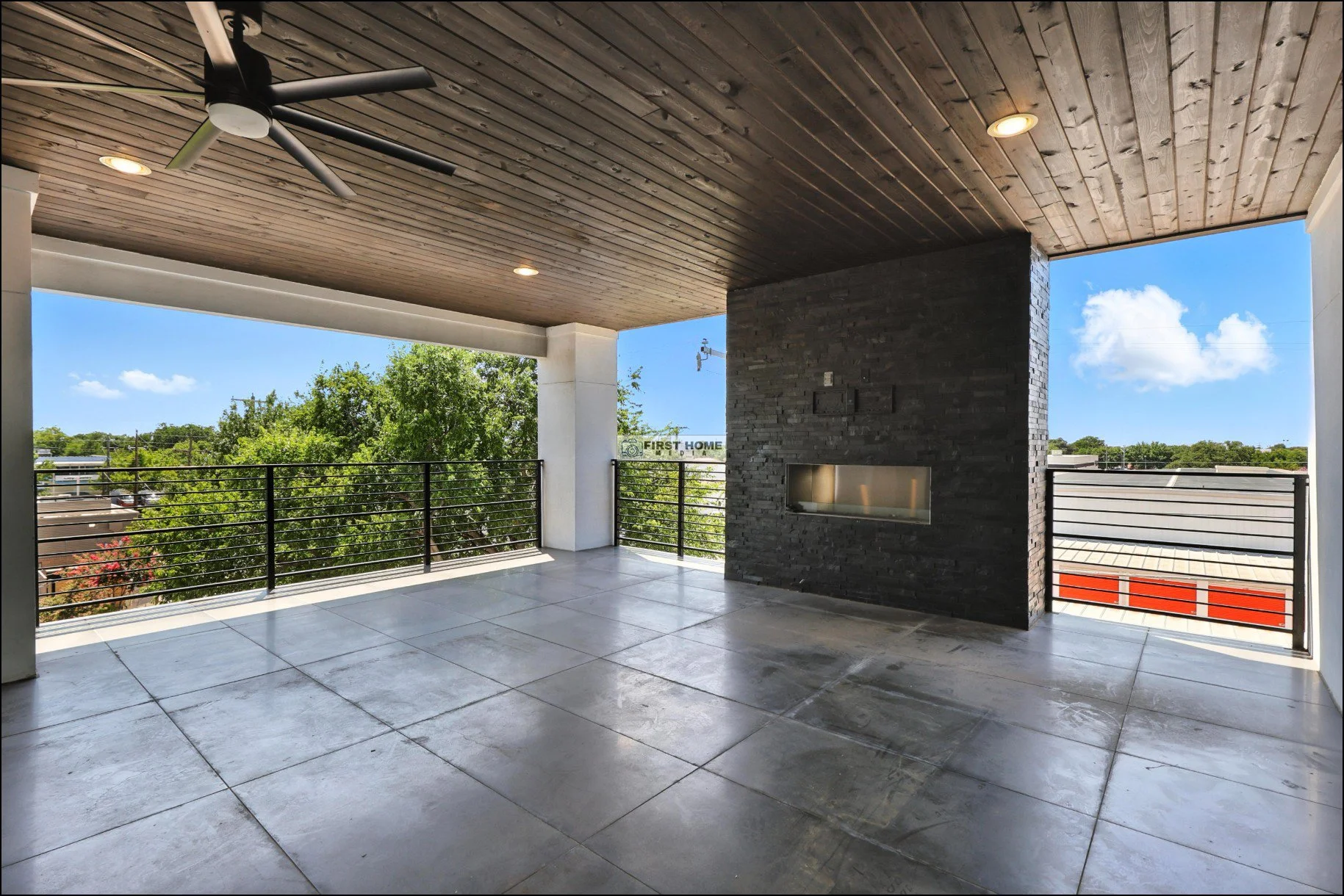 Empty outdoor balcony with black metal railing, wooden ceiling, tiled floor, and a built-in fireplace, overlooking trees and nearby buildings under a partly cloudy sky.