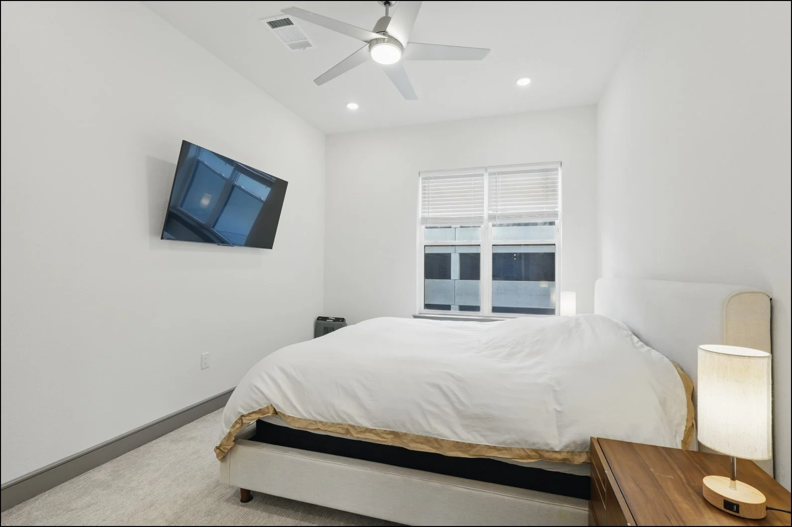 Modern bedroom with a mounted flat-screen TV, white bed, ceiling fan, nightstand, and window with blinds.