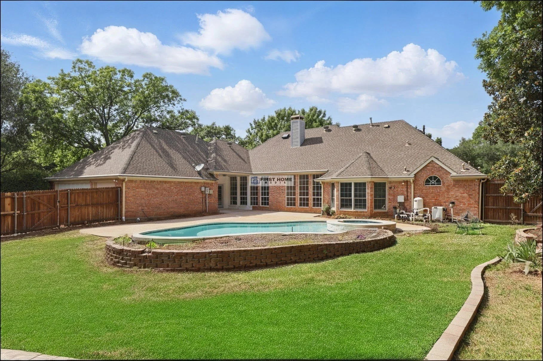 Backyard of a brick house with a swimming pool, green lawn, trees, and a clear blue sky with white clouds.