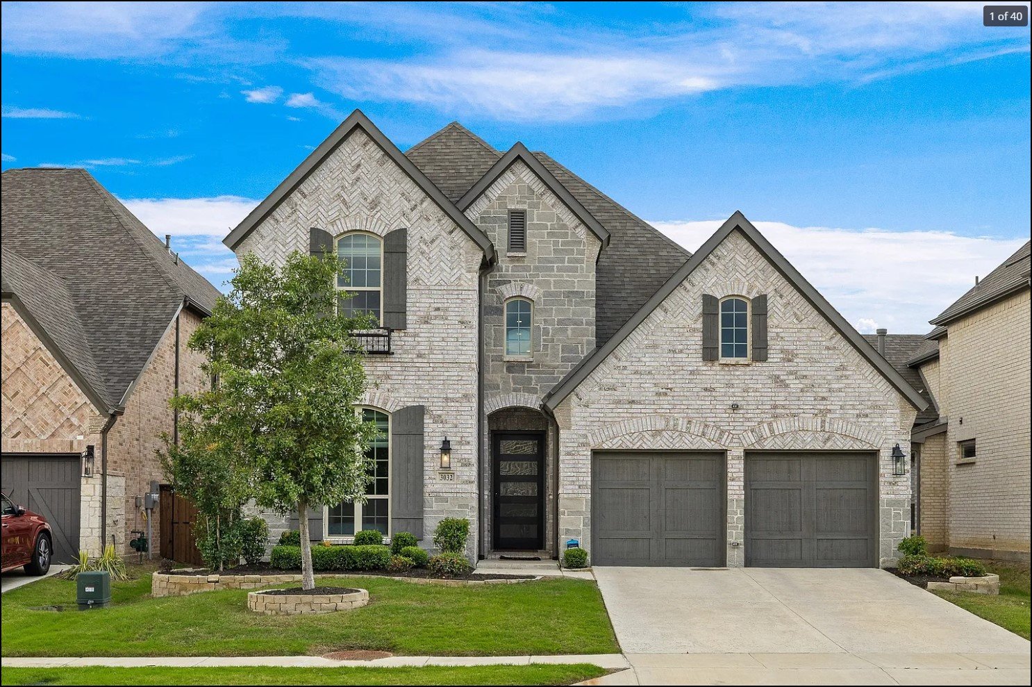 Two-story modern brick house with gray roof, shutters, and double garage. A tree and landscaped lawn in front.