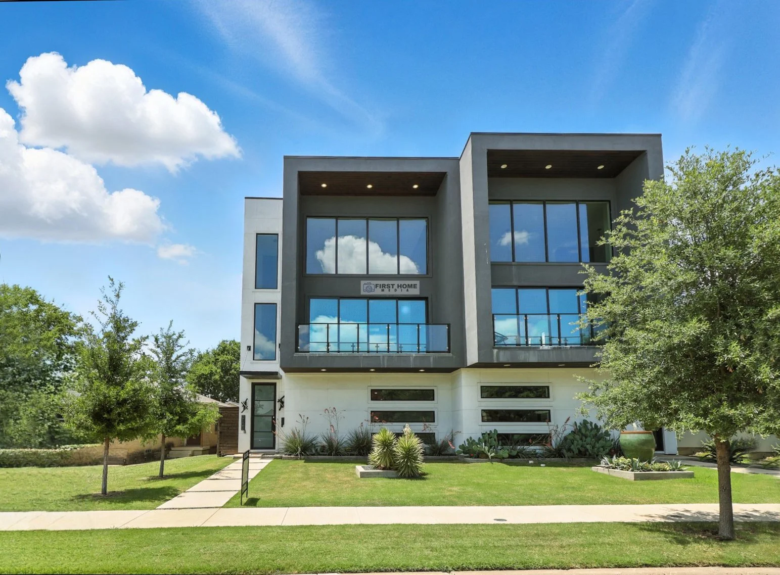 Modern multi-story house with large glass windows, a well-maintained lawn, and trees under a bright blue sky with scattered clouds.