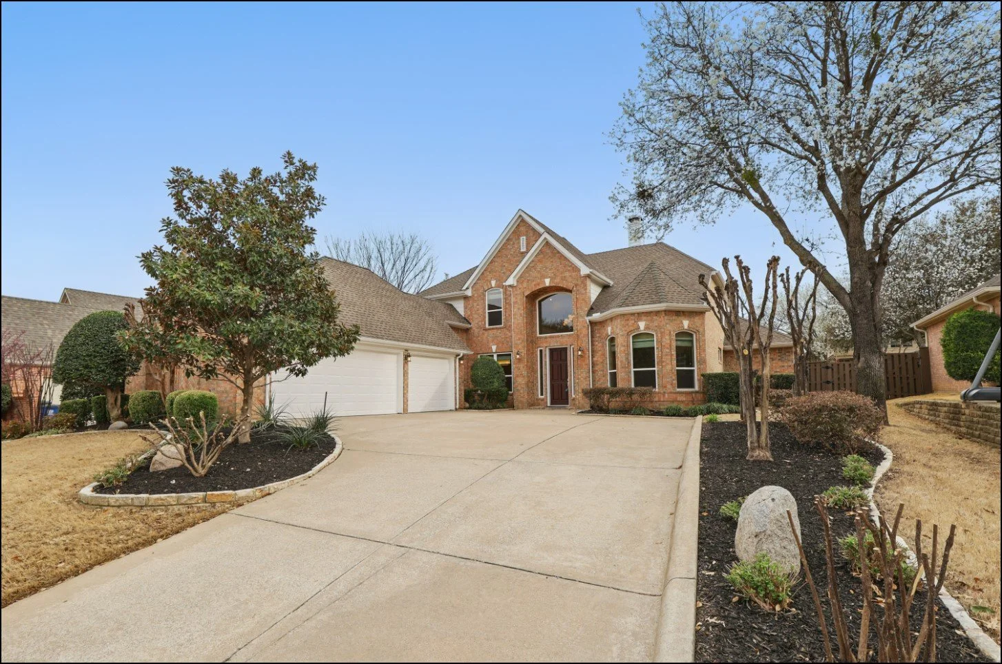 Two-story brick house with a three-car garage, large driveway, manicured lawn, and mature trees.