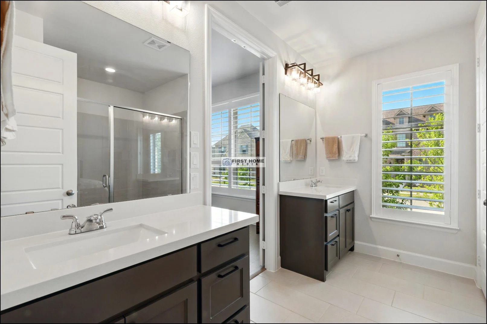Bathroom with two separate vanities, large mirror, beige and white towels, window with white blinds, glass-enclosed shower, and a modern light fixture.