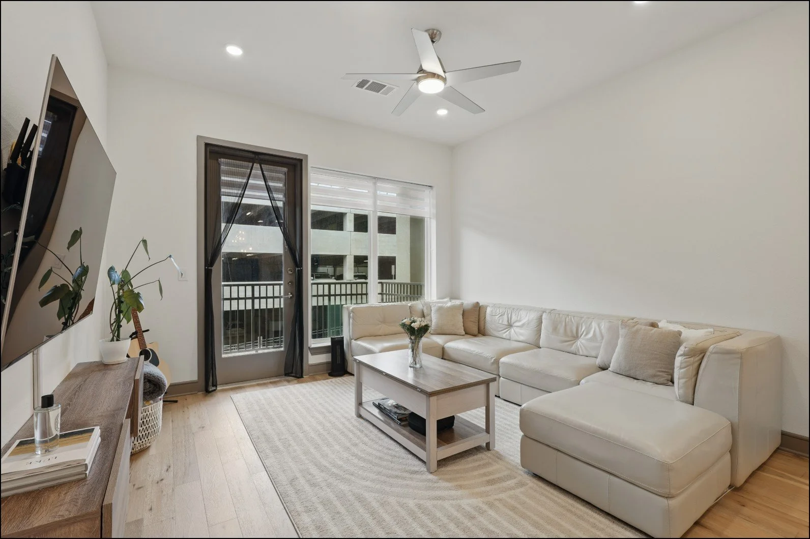 Modern living room with beige sectional sofa, wooden coffee table, ceiling fan, and large windows with a balcony view. A TV is mounted on the wall, and a guitar is placed near a potted plant.