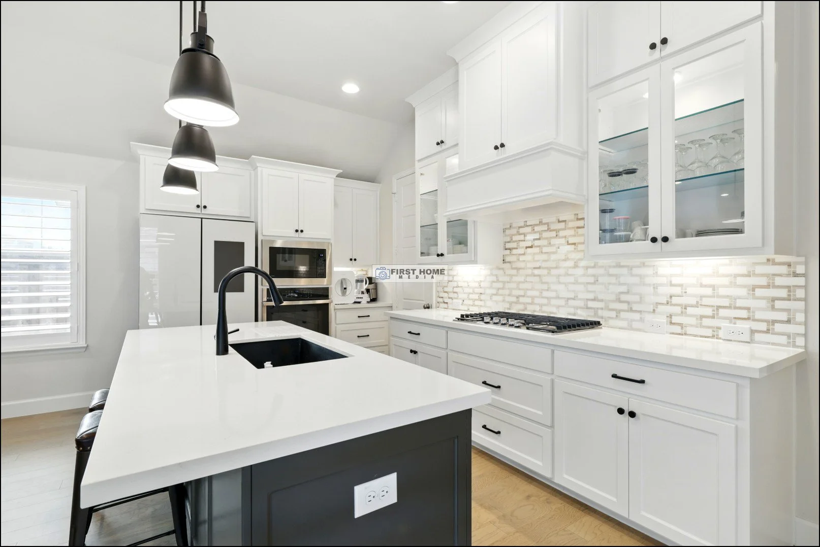 Modern kitchen with white cabinets, black hardware, a black island with an integrated sink, pendant lights, stainless steel appliances, a white tiled backsplash, and hardwood floors.