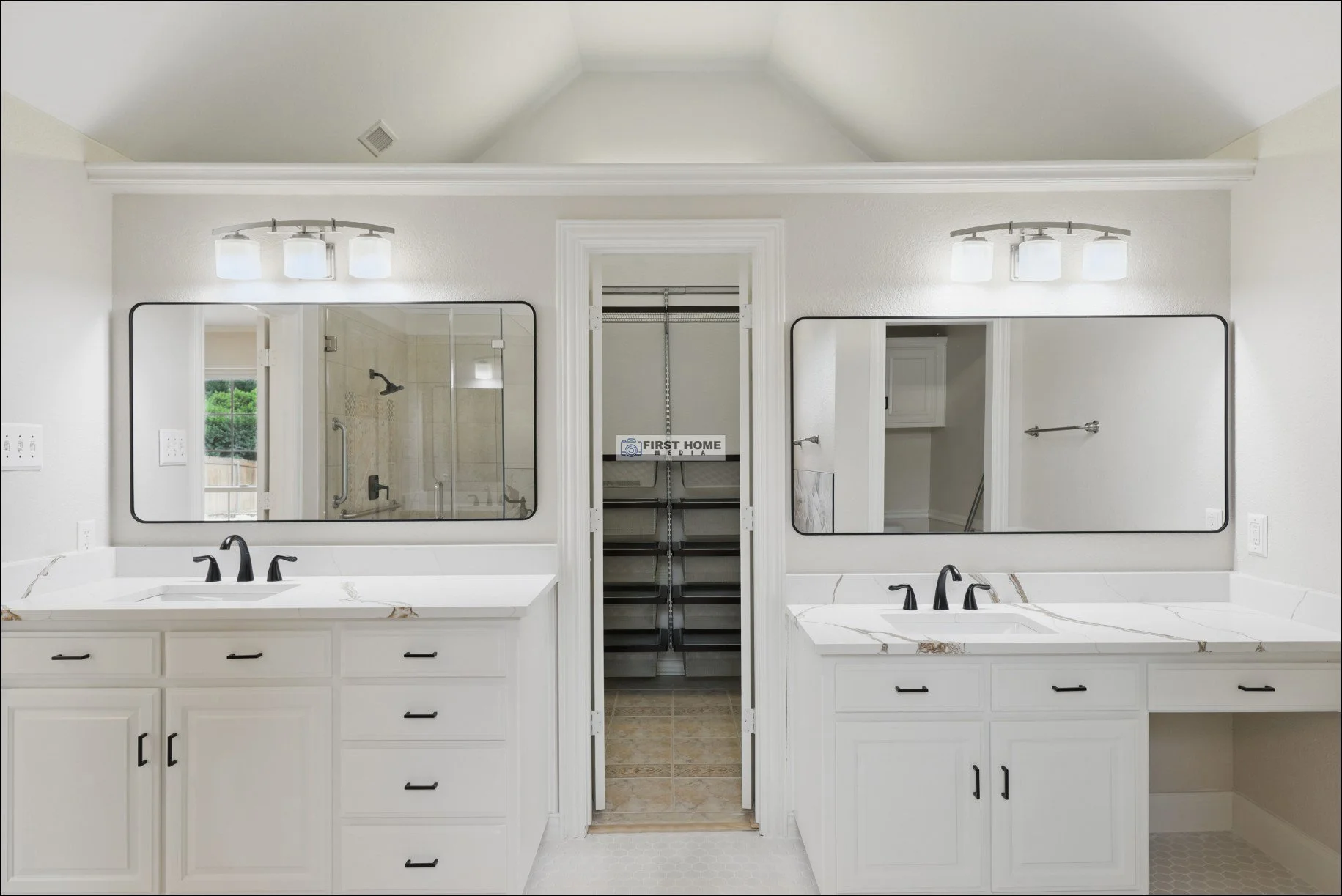 Double vanity bathroom with white cabinets, marble countertops, black fixtures, large mirrors, wall-mounted lights, and a walk-in closet between the sinks.