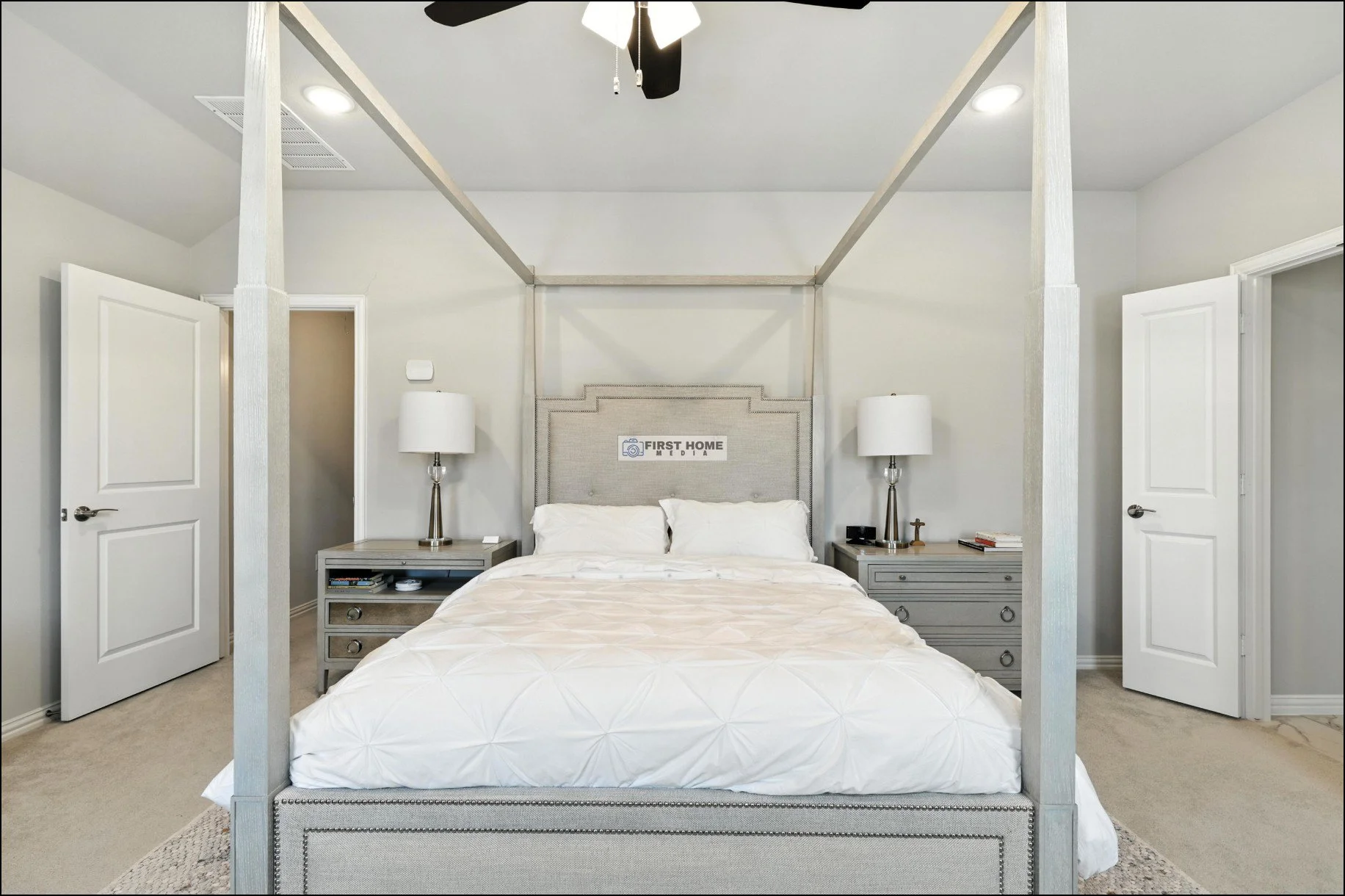 A bedroom with a four-poster bed, beige headboard, white bedding, and two matching gray nightstands with lamps. Two doors open into the room, a ceiling fan hangs above, and the walls are painted light gray.