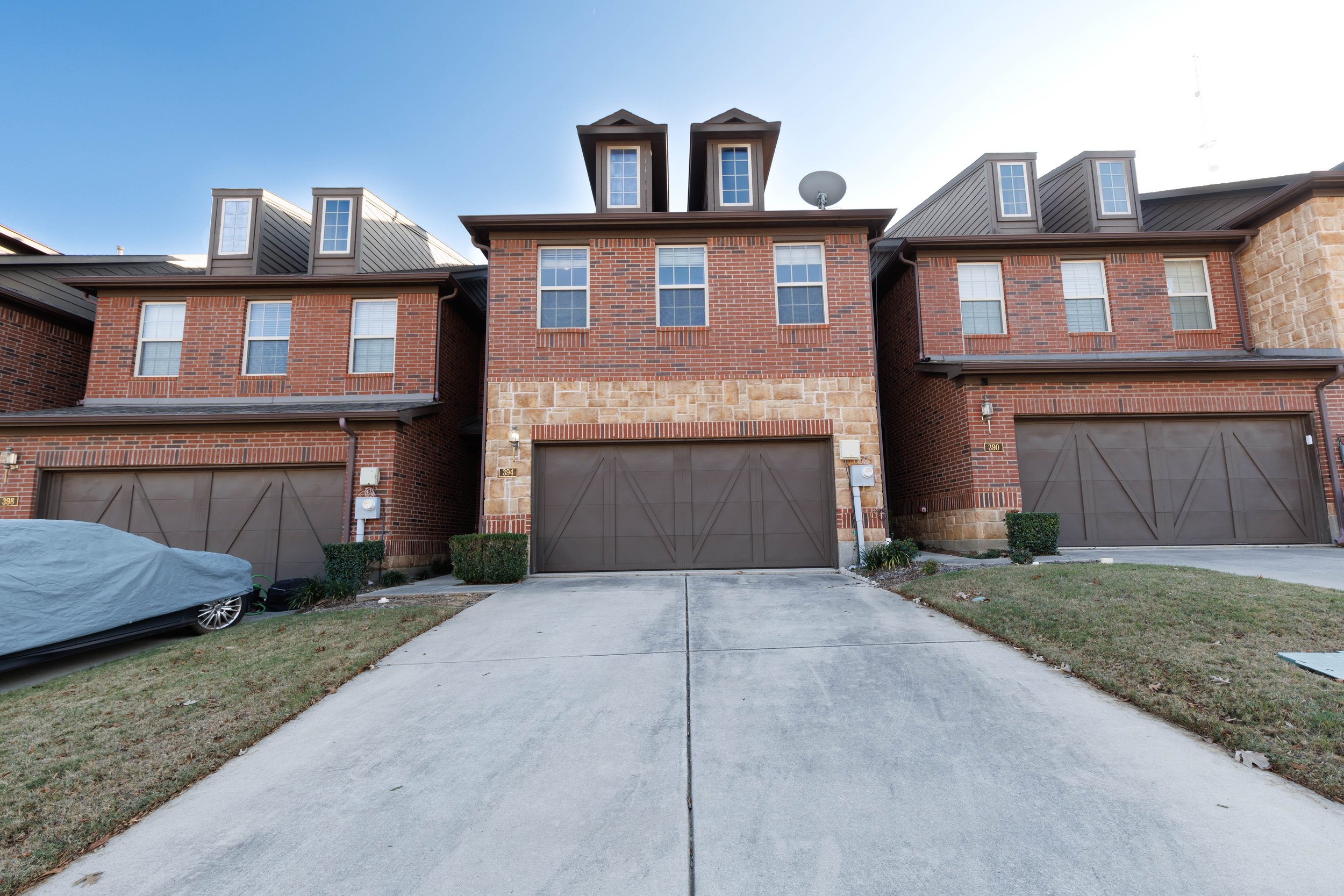 Row of three-story brick townhouses with pitched roofs, double garage doors, and a covered car in the driveway.